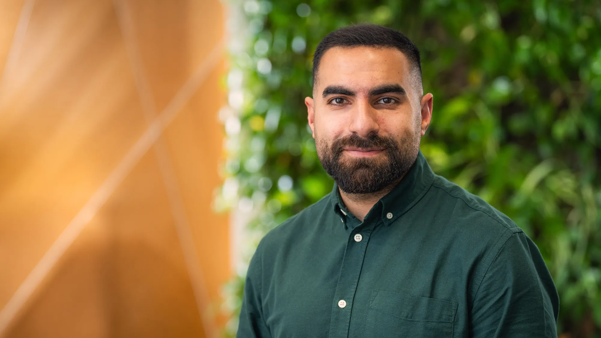 Person wearing a dark green button-up shirt stands in front of a background with green foliage and a wooden wall featuring diagonal lines.