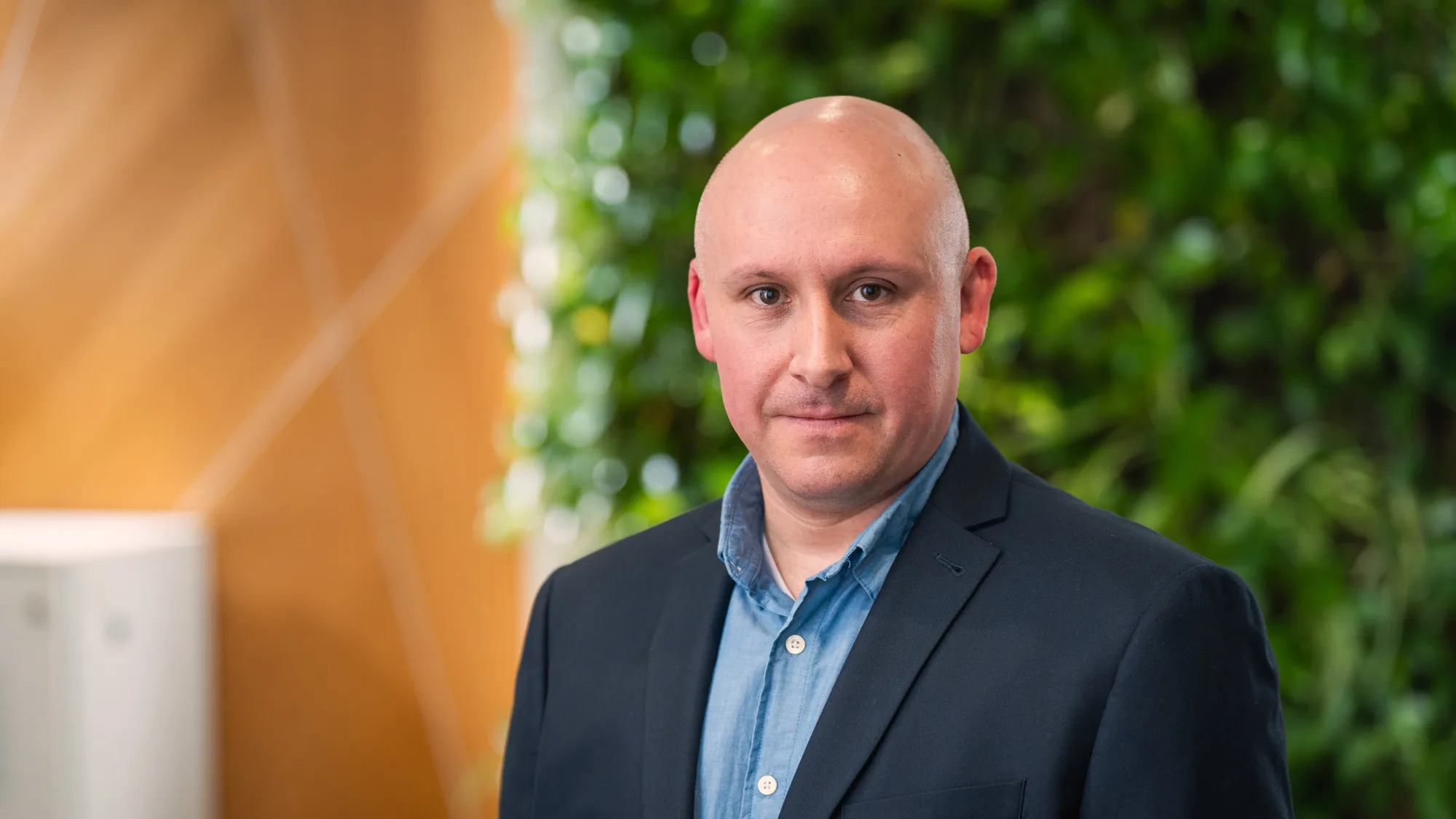 Tony looking to camera in an open collar shirt and suit jacket in front of a living wall