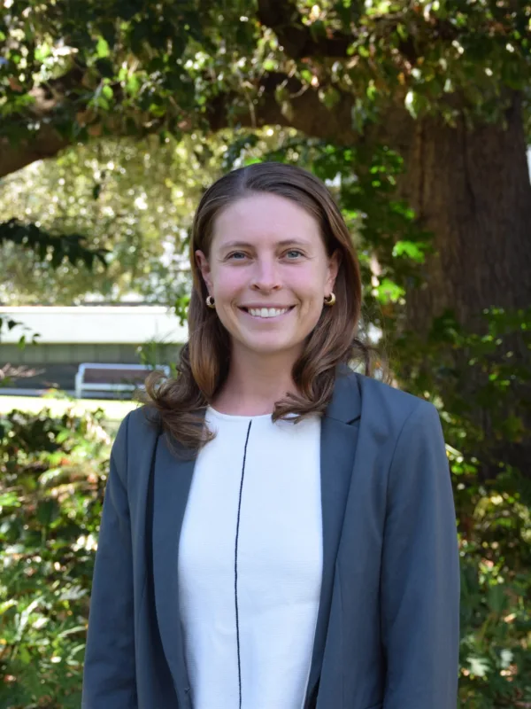 Anna Gannett wearing a light-colored top and a gray blazer standing outdoors in front of large tree and greenery, with benches and a building partially visible in the background.