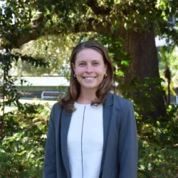 Anna Gannett wearing a light-colored top and a gray blazer standing outdoors in front of large tree and greenery, with benches and a building partially visible in the background.