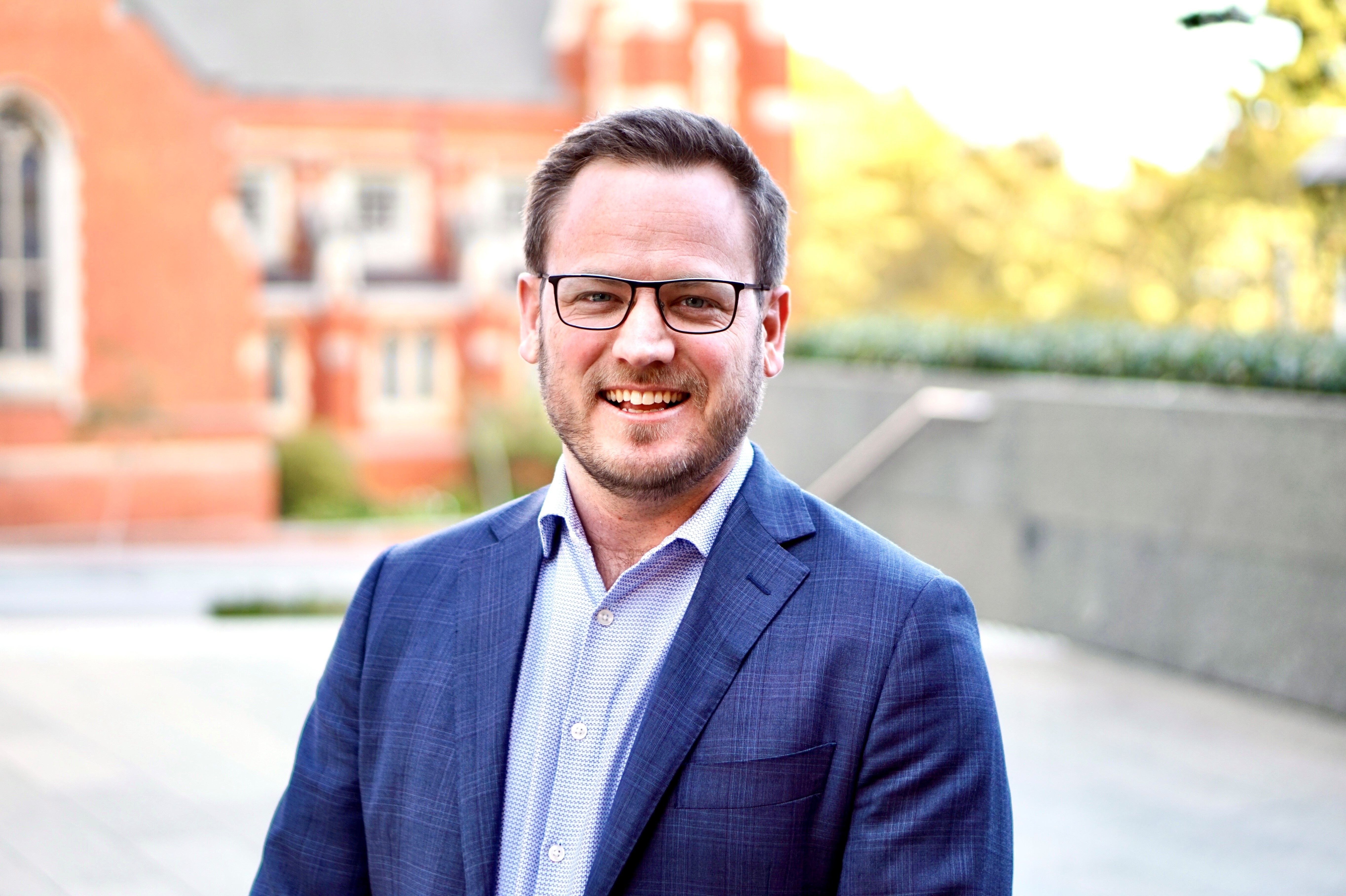 Head and sholders shot of Tim in dark blue blazer and light blue shirt wearing glasses against a bright blurred out background