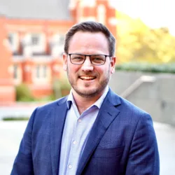 Head and sholders shot of Tim in dark blue blazer and light blue shirt wearing glasses against a bright blurred out background