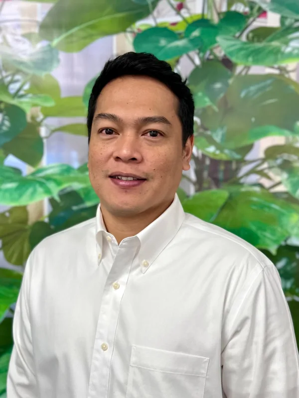 Jayson Landayan wearing a white button-down shirt with a front pocket, standing in front of a backdrop featuring large green leaves and plants.