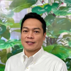 Jayson Landayan wearing a white button-down shirt with a front pocket, standing in front of a backdrop featuring large green leaves and plants.