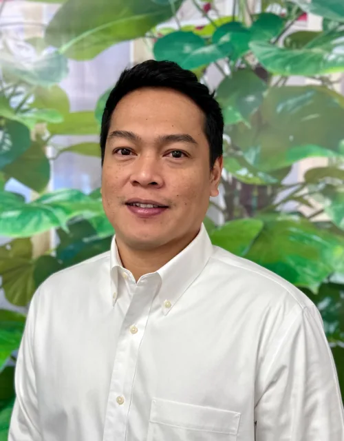 Jayson Landayan wearing a white button-down shirt with a front pocket, standing in front of a backdrop featuring large green leaves and plants.