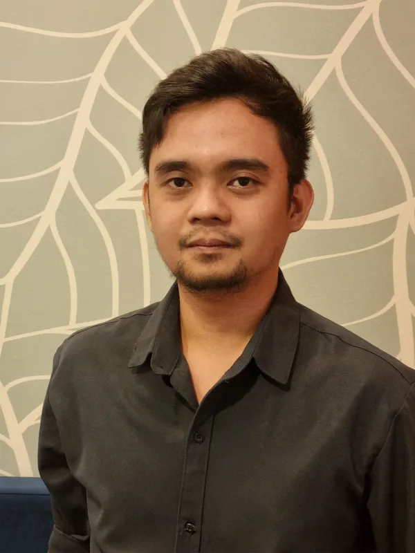 Person standing indoors wearing a dark long-sleeve button-up shirt, photographed from the waist up. Behind is a light wall with a white leaf pattern and a blue upholstered bench along the lower edge.