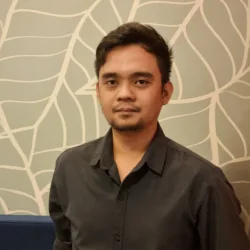 Person standing indoors wearing a dark long-sleeve button-up shirt, photographed from the waist up. Behind is a light wall with a white leaf pattern and a blue upholstered bench along the lower edge.