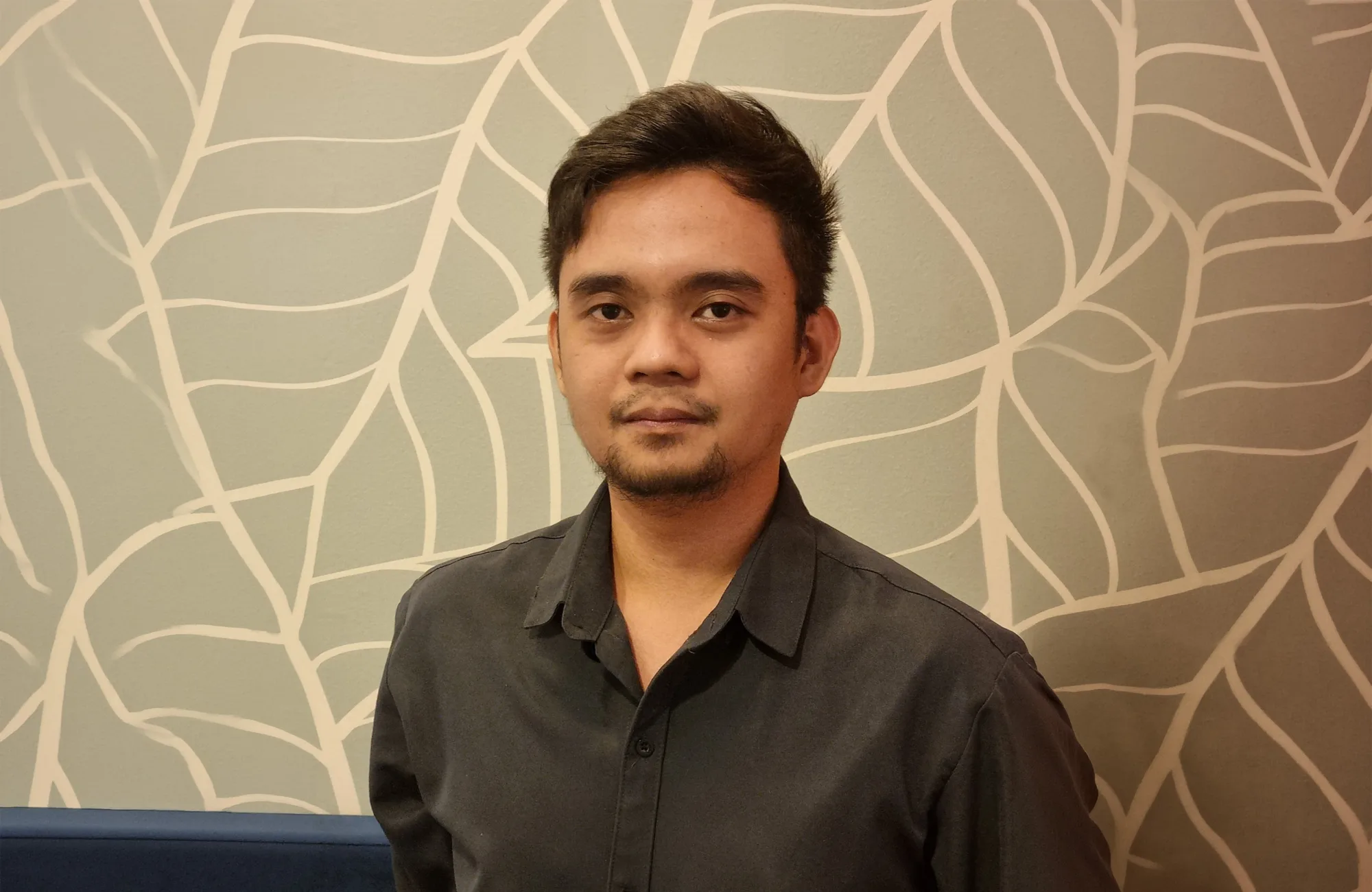 Person standing indoors wearing a dark long-sleeve button-up shirt, photographed from the waist up. Behind is a light wall with a white leaf pattern and a blue upholstered bench along the lower edge.