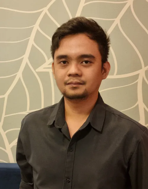Person standing indoors wearing a dark long-sleeve button-up shirt, photographed from the waist up. Behind is a light wall with a white leaf pattern and a blue upholstered bench along the lower edge.