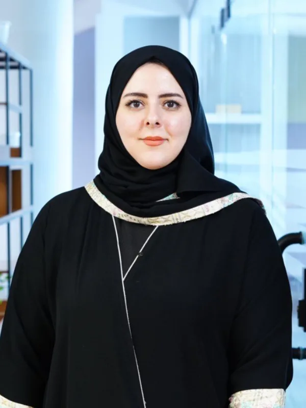 A person wearing a black head covering and black outfit stands facing the camera in a modern office corridor. Shelving with plants lines the left side, and a glass door and bright interior are visible in the background.
