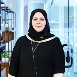 A person wearing a black head covering and black outfit stands facing the camera in a modern office corridor. Shelving with plants lines the left side, and a glass door and bright interior are visible in the background.