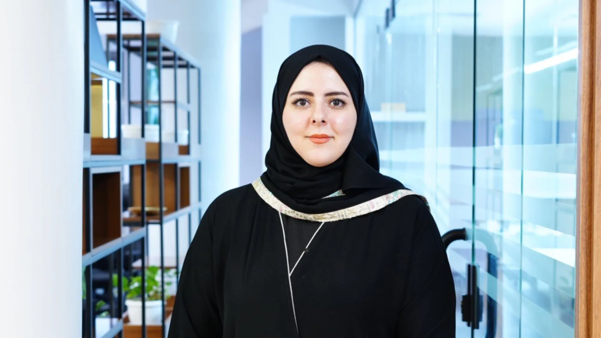 A person wearing a black head covering and black outfit stands facing the camera in a modern office corridor. Shelving with plants lines the left side, and a glass door and bright interior are visible in the background.