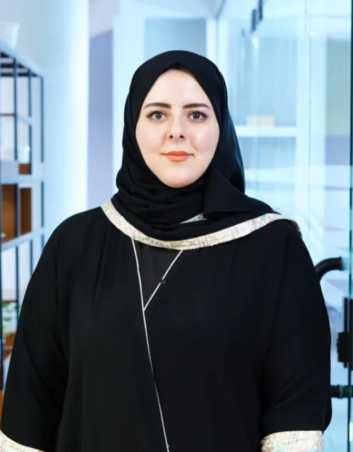 A person wearing a black head covering and black outfit stands facing the camera in a modern office corridor. Shelving with plants lines the left side, and a glass door and bright interior are visible in the background.