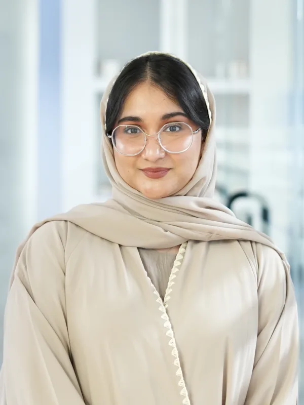 Ayesha Ahmed in light-colored garment with head covering standing indoors; background includes shelves with items, plants, and glass partitions in a modern, organized setting.