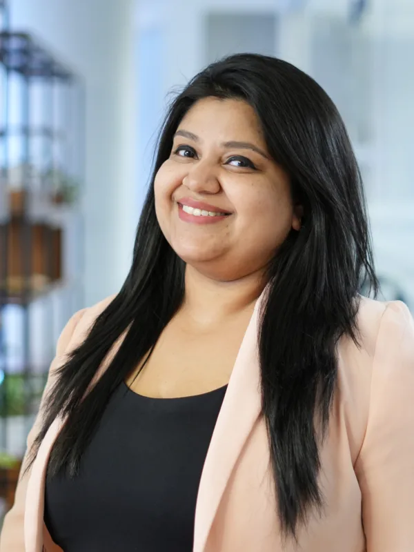 A person with long dark hair stands in an office setting, wearing a light-coloured blazer over a black top. Behind them are shelves with plants, glass walls, doors, and office furniture.