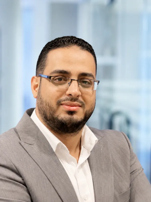 Portrait of a person wearing rectangular glasses, a light grey blazer, and a white shirt, facing the camera. Background shows a softly blurred modern office interior with shelves and glass partitions.
