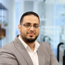 Portrait of a person wearing rectangular glasses, a light grey blazer, and a white shirt, facing the camera. Background shows a softly blurred modern office interior with shelves and glass partitions.