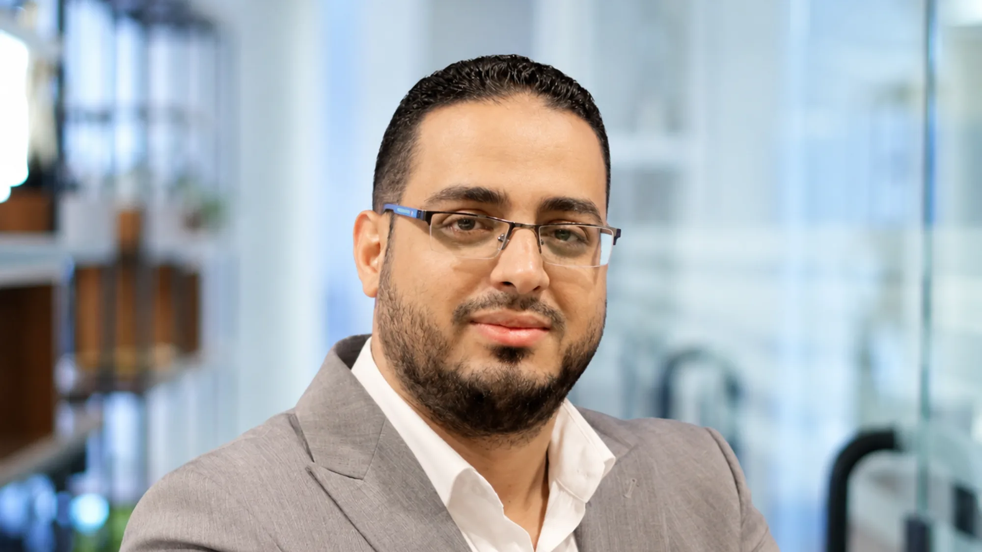 Portrait of a person wearing rectangular glasses, a light grey blazer, and a white shirt, facing the camera. Background shows a softly blurred modern office interior with shelves and glass partitions.