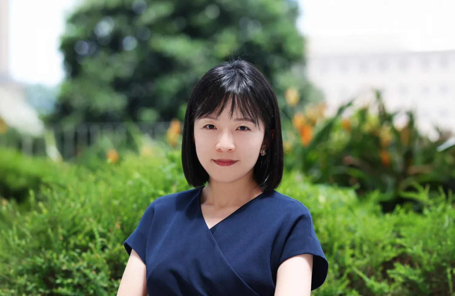Portrait of a person with straight, chin-length dark hair wearing a navy short-sleeved top, standing outdoors in front of green shrubs and flowers, with a softly blurred tree and building in the background under daylight.