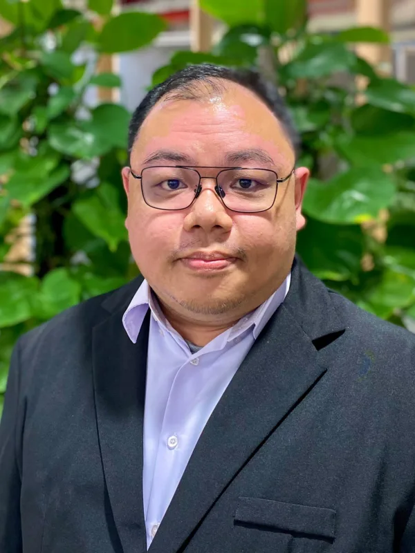 Man wearing a dark suit jacket and light dress shirt stands in front of a wooden slat wall with green leafy plants. Red pipes and industrial elements are visible in the indoor background.