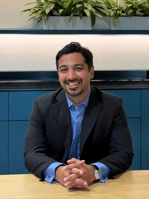 A person wearing a dark suit jacket and light blue shirt sits at a wooden table with hands folded. Behind them is a blue cabinet and a row of green plants on a shelf.