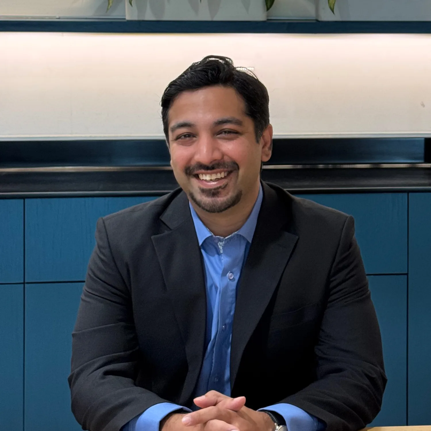 A person wearing a dark suit jacket and light blue shirt sits at a wooden table with hands folded. Behind them is a blue cabinet and a row of green plants on a shelf.