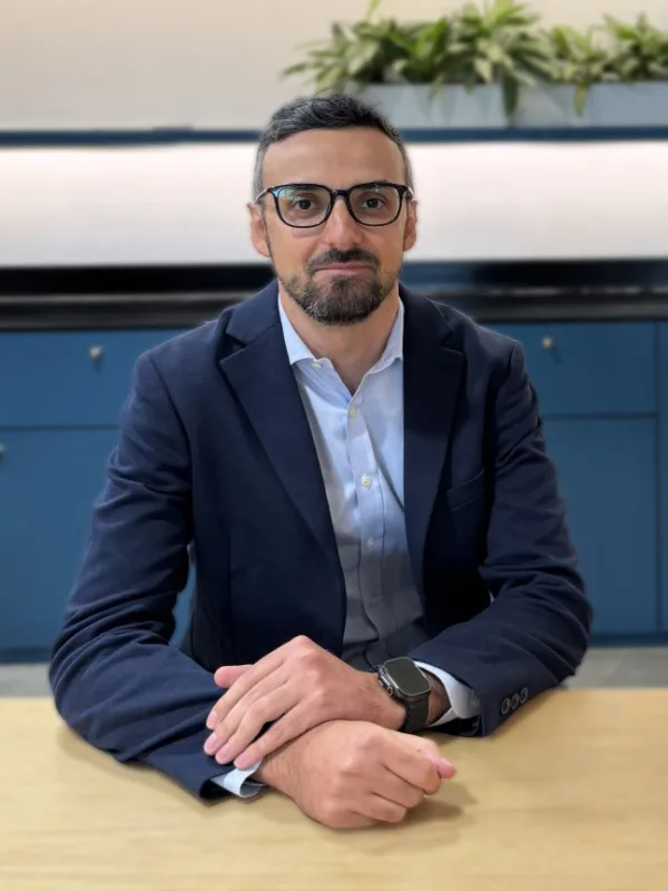 A person in a navy blazer and light blue shirt sits at a wooden table with hands folded. They wear a smartwatch on their left wrist. Behind them is a blue cabinet, wall sockets, and a row of green plants.