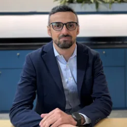 A person in a navy blazer and light blue shirt sits at a wooden table with hands folded. They wear a smartwatch on their left wrist. Behind them is a blue cabinet, wall sockets, and a row of green plants.