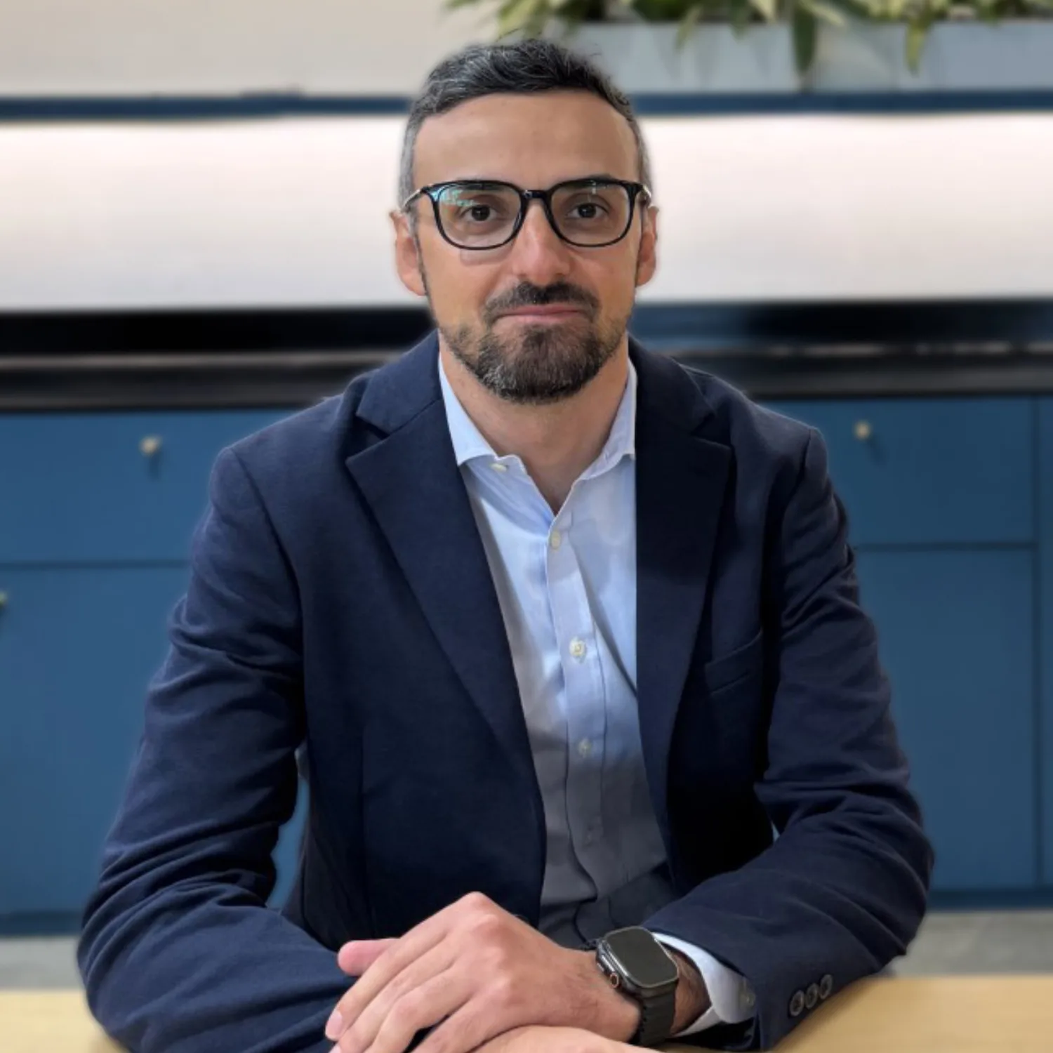 A person in a navy blazer and light blue shirt sits at a wooden table with hands folded. They wear a smartwatch on their left wrist. Behind them is a blue cabinet, wall sockets, and a row of green plants.