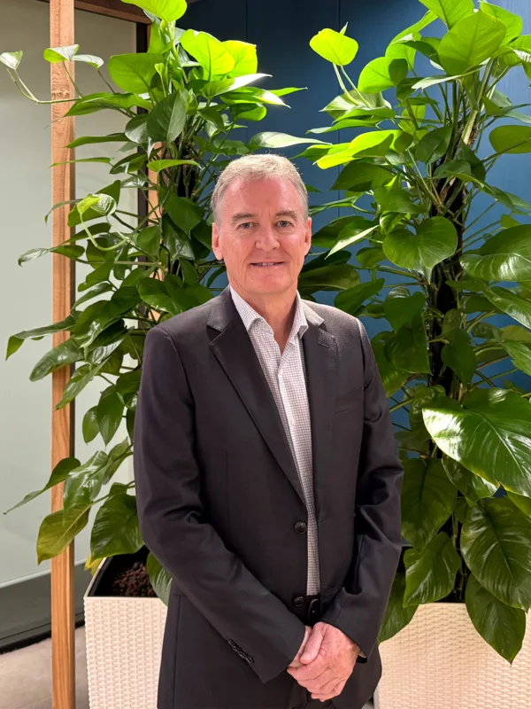 A person wearing a dark suit jacket and light shirt stands indoors in front of tall green plants, with glass panels and wooden frames behind.