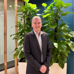 A person wearing a dark suit jacket and light shirt stands indoors in front of tall green plants, with glass panels and wooden frames behind.