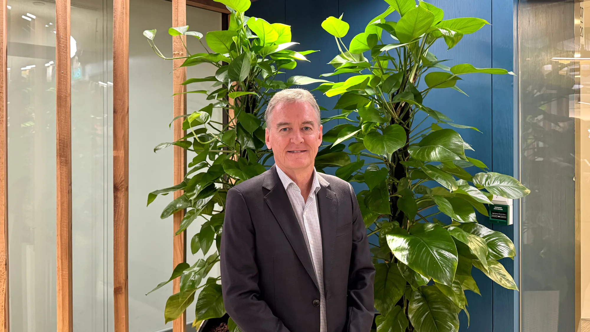 A person wearing a dark suit jacket and light shirt stands indoors in front of tall green plants, with glass panels and wooden frames behind.