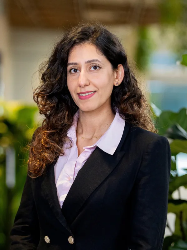 Portrait of a person with long, curly dark hair, wearing a black blazer over a light pink collared shirt, facing the camera. Background shows soft-focus greenery and warm indoor lighting, with no visible text.