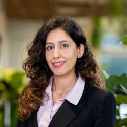 Portrait of a person with long, curly dark hair, wearing a black blazer over a light pink collared shirt, facing the camera. Background shows soft-focus greenery and warm indoor lighting, with no visible text.
