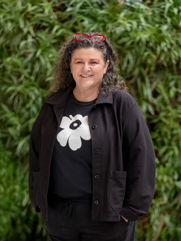 Bettina in dark clothing in front of a green hedge with red glasses on her head smiling to camera