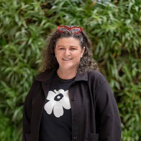 Bettina in dark clothing in front of a green hedge with red glasses on her head smiling to camera