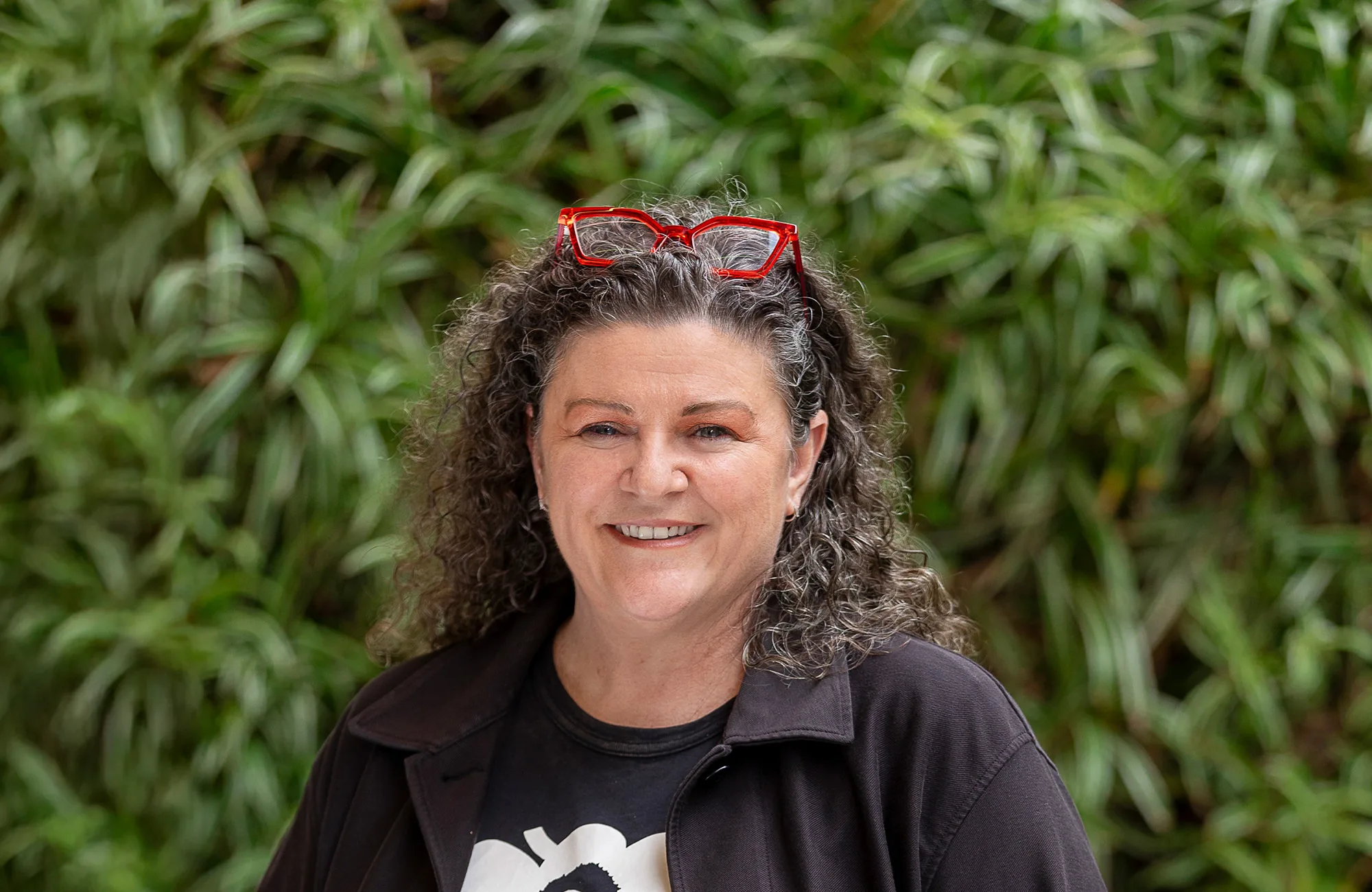 Bettina in dark clothing in front of a green hedge with red glasses on her head smiling to camera