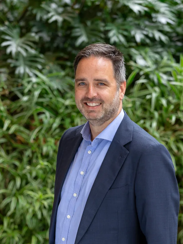 Julian, stood shoulder forward in a dark blue suit jacket and pale blue shite, smiling in front of a green hedge.