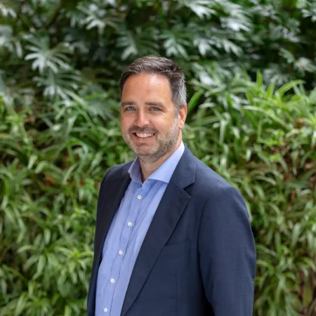 Julian, stood shoulder forward in a dark blue suit jacket and pale blue shite, smiling in front of a green hedge.