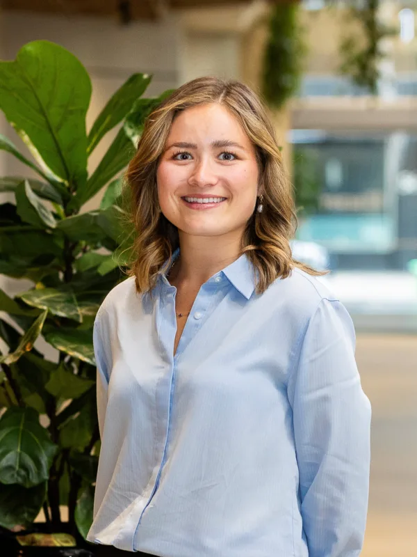 Portrait of a person standing indoors beside a large leafy plant, wearing a light blue button-down shirt, facing the camera. Background shows a bright office space with wooden ceiling details.