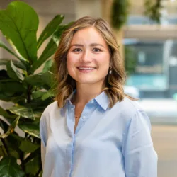 Portrait of a person standing indoors beside a large leafy plant, wearing a light blue button-down shirt, facing the camera. Background shows a bright office space with wooden ceiling details.