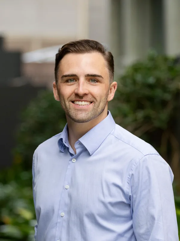 Portrait of a person standing outdoors in a walkway with greenery, wearing a light blue button-down shirt, facing camera; background shows plants and blurred building elements.