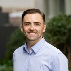 Portrait of a person standing outdoors in a walkway with greenery, wearing a light blue button-down shirt, facing camera; background shows plants and blurred building elements.