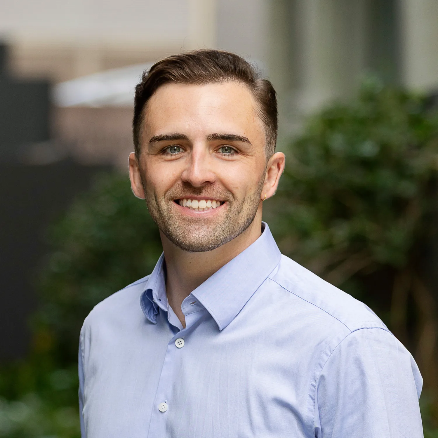 Portrait of a person standing outdoors in a walkway with greenery, wearing a light blue button-down shirt, facing camera; background shows plants and blurred building elements.