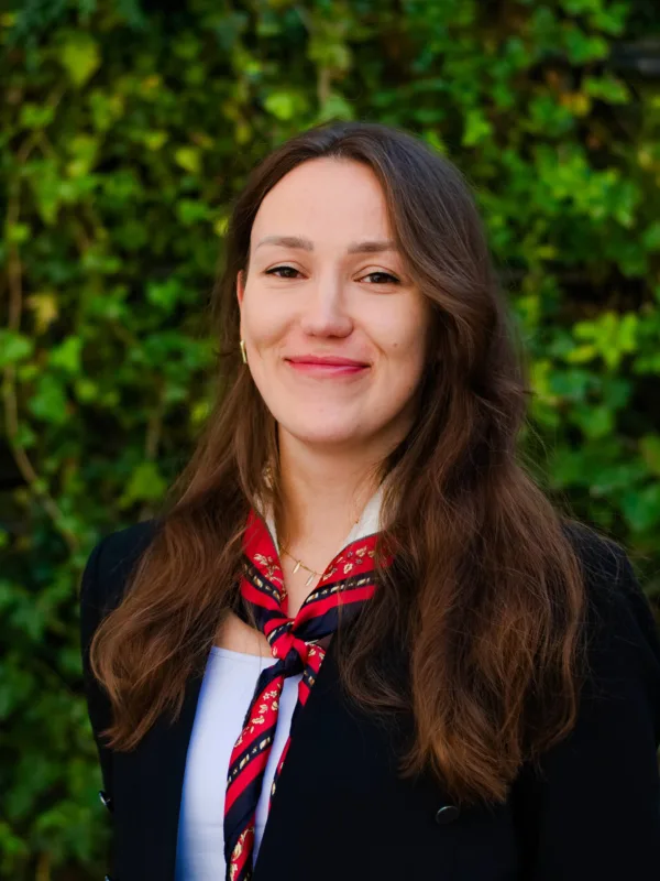 A person with long brown hair is standing outdoors in front of a dense green leafy background. They are wearing a black jacket over a white top and a red patterned scarf tied around the neck.