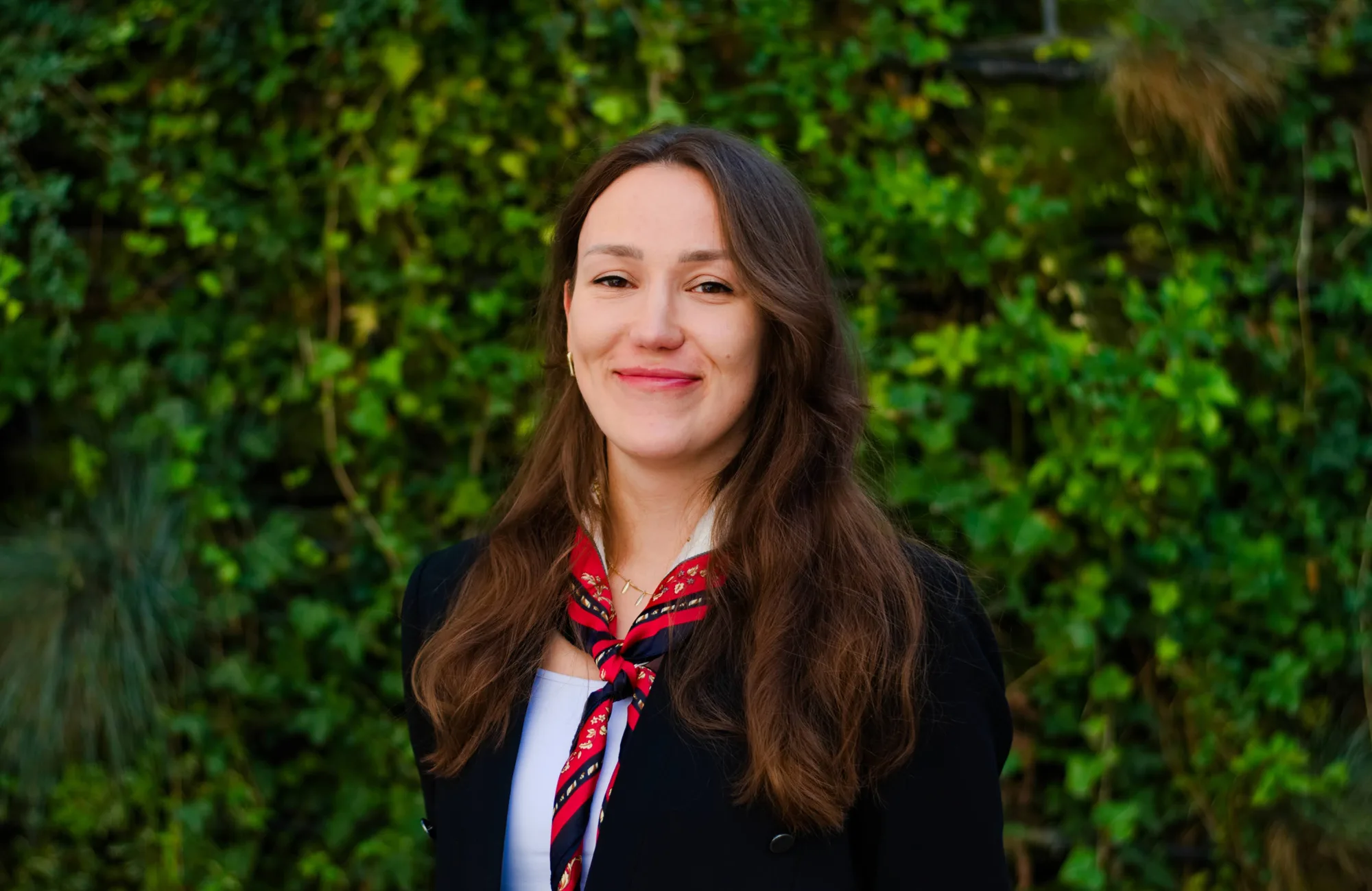 A person with long brown hair is standing outdoors in front of a dense green leafy background. They are wearing a black jacket over a white top and a red patterned scarf tied around the neck.