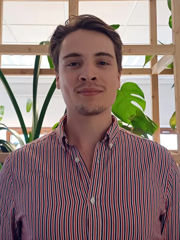 Maciej in a striped open collar shirt looking to camera in front of an open wooden shelving unt with green plants.