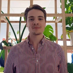 Maciej in a striped open collar shirt looking to camera in front of an open wooden shelving unt with green plants.
