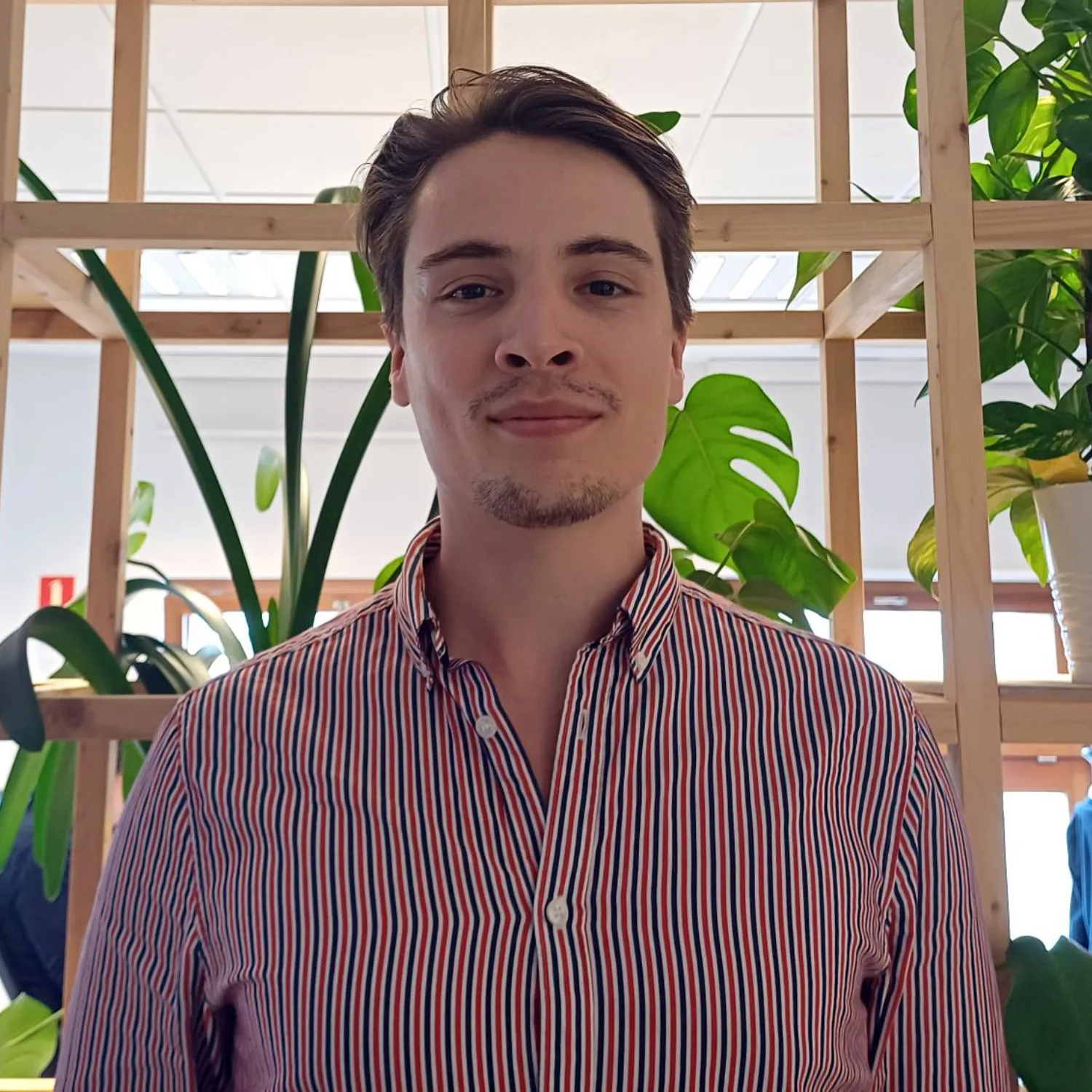 Maciej in a striped open collar shirt looking to camera in front of an open wooden shelving unt with green plants.