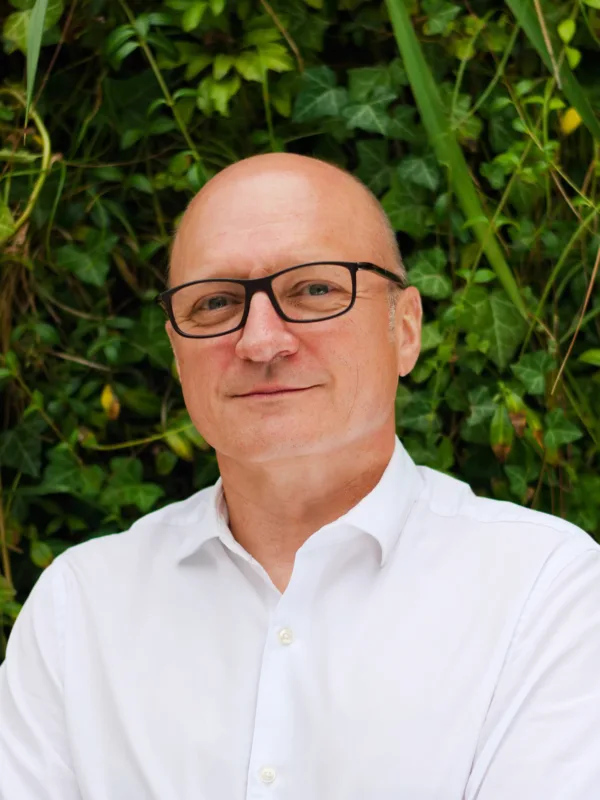 Slawomir standing in front of a camera wearing white shirt with plants on the background.